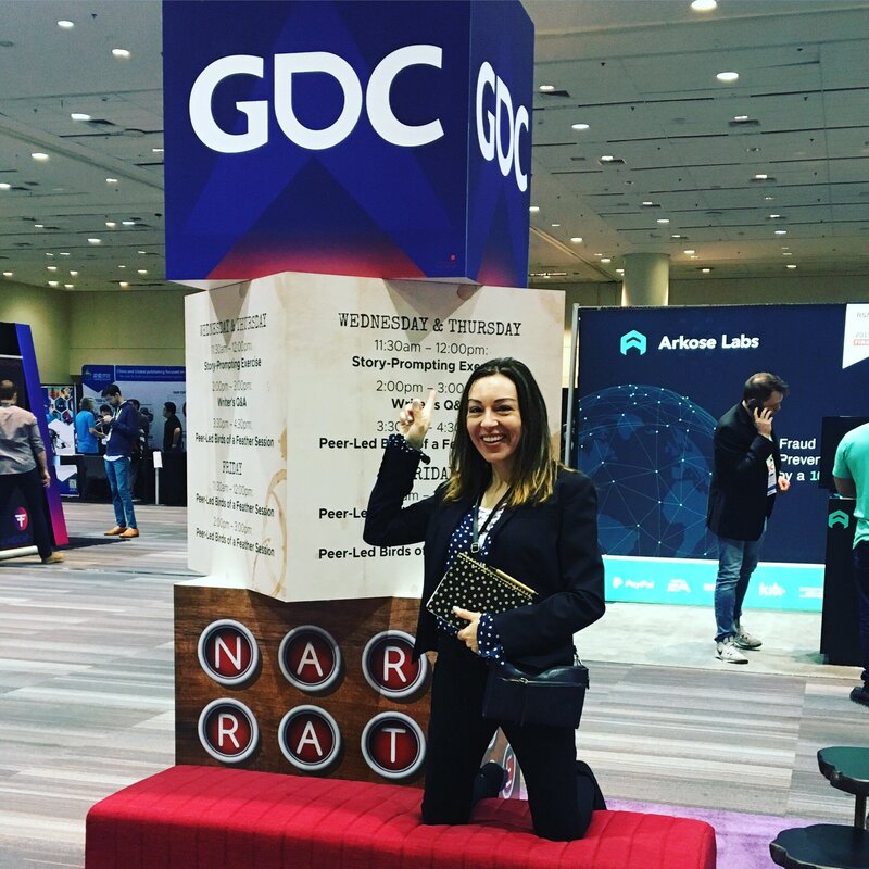 A woman (Nelia) smiles and points at a sign under a large GDC cube at a convention.