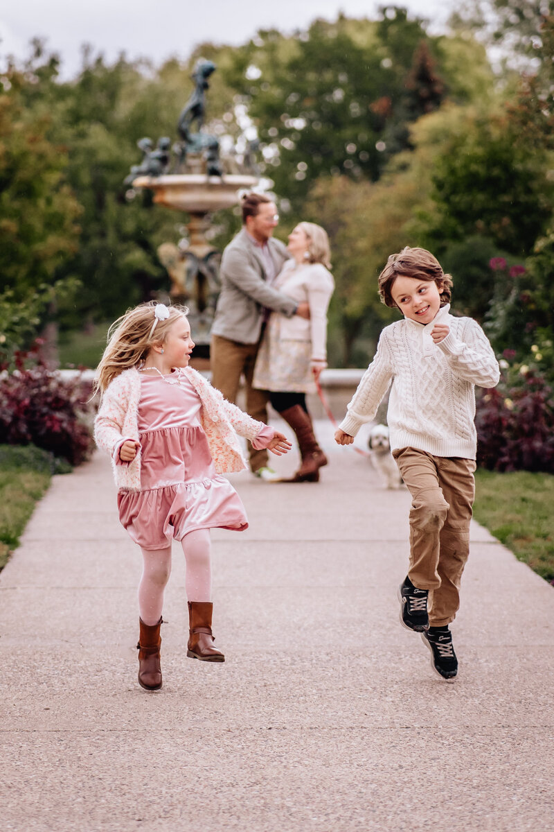 Two children run joyfully down a path in a park, while their parents stand in the background, embracing and watching them with smiles.