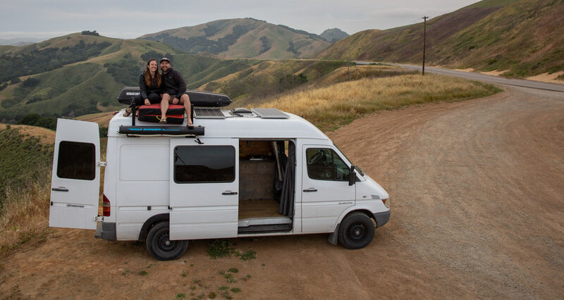 Becky and Brian sit on top of their van  with the rolling hills of Central Coast California behind them.