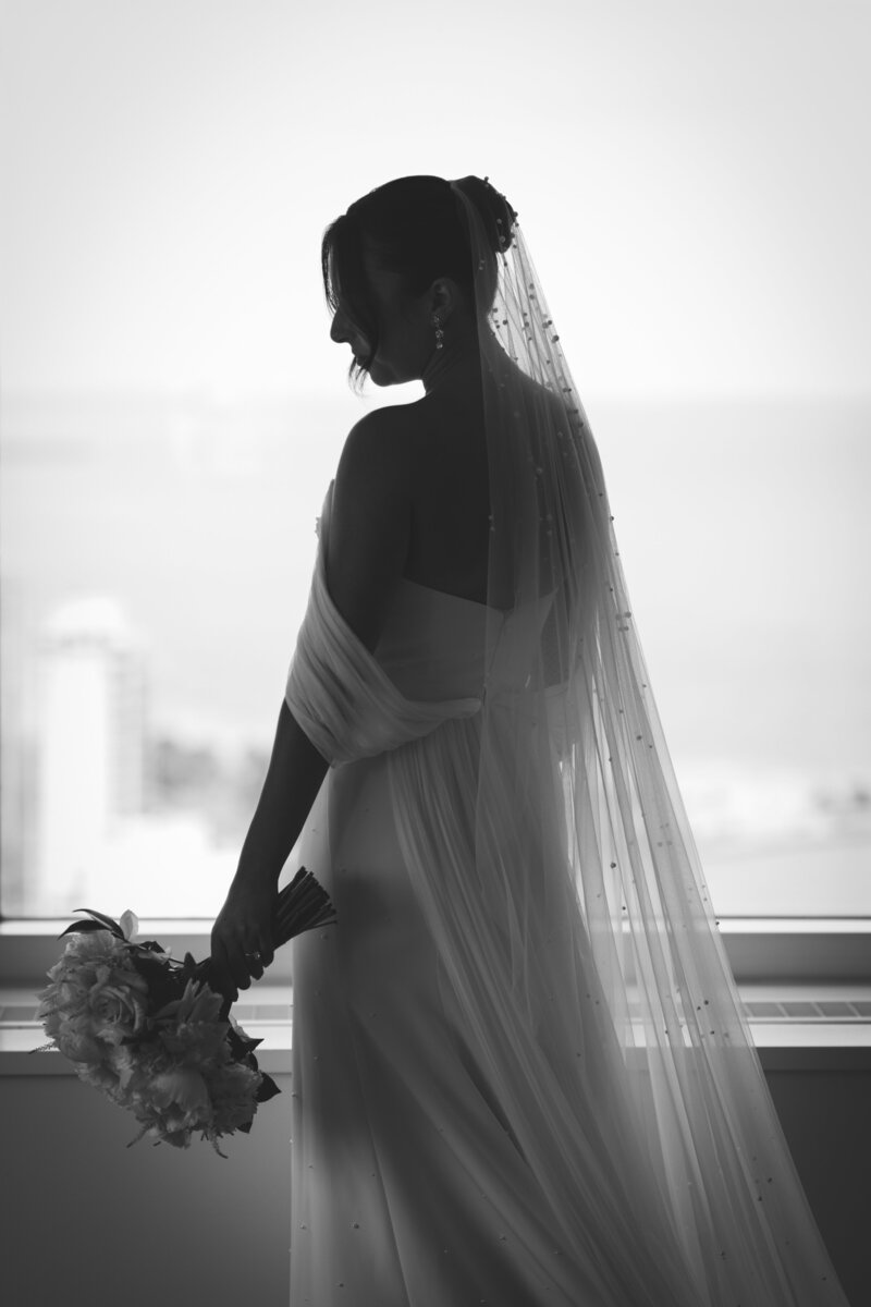 A silhouette of a bride holding her bouquet, standing by a window with a cityscape in the background, during her Four Seasons Chicago wedding planned by Savoir Fête.