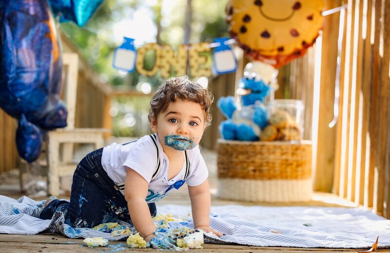 A happy baby boy in a football outfit sits on a blanket with a football cake, during a fun cake smash photo session.