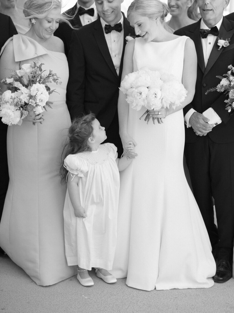 Bride looks down at her flower girl as wedding parties pose for portraits