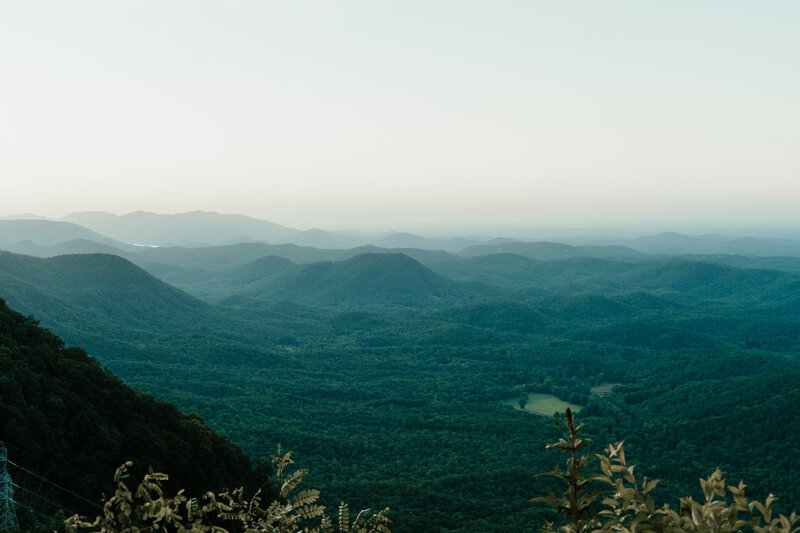 A mountaintop view in North Carolina at golden hour, with layered peaks fading into soft blues. A summit marker in the foreground hints at love rooted in adventure.