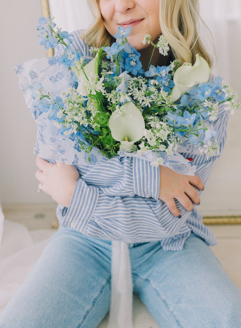 Close-up of a blue and white wildflower bouquet wrapped in floral paper, held by a woman in a blue striped shirt. Branding photo with soft light and natural expression. Greenville South Carolina portrait photographer – Dani Lacey Photographs.