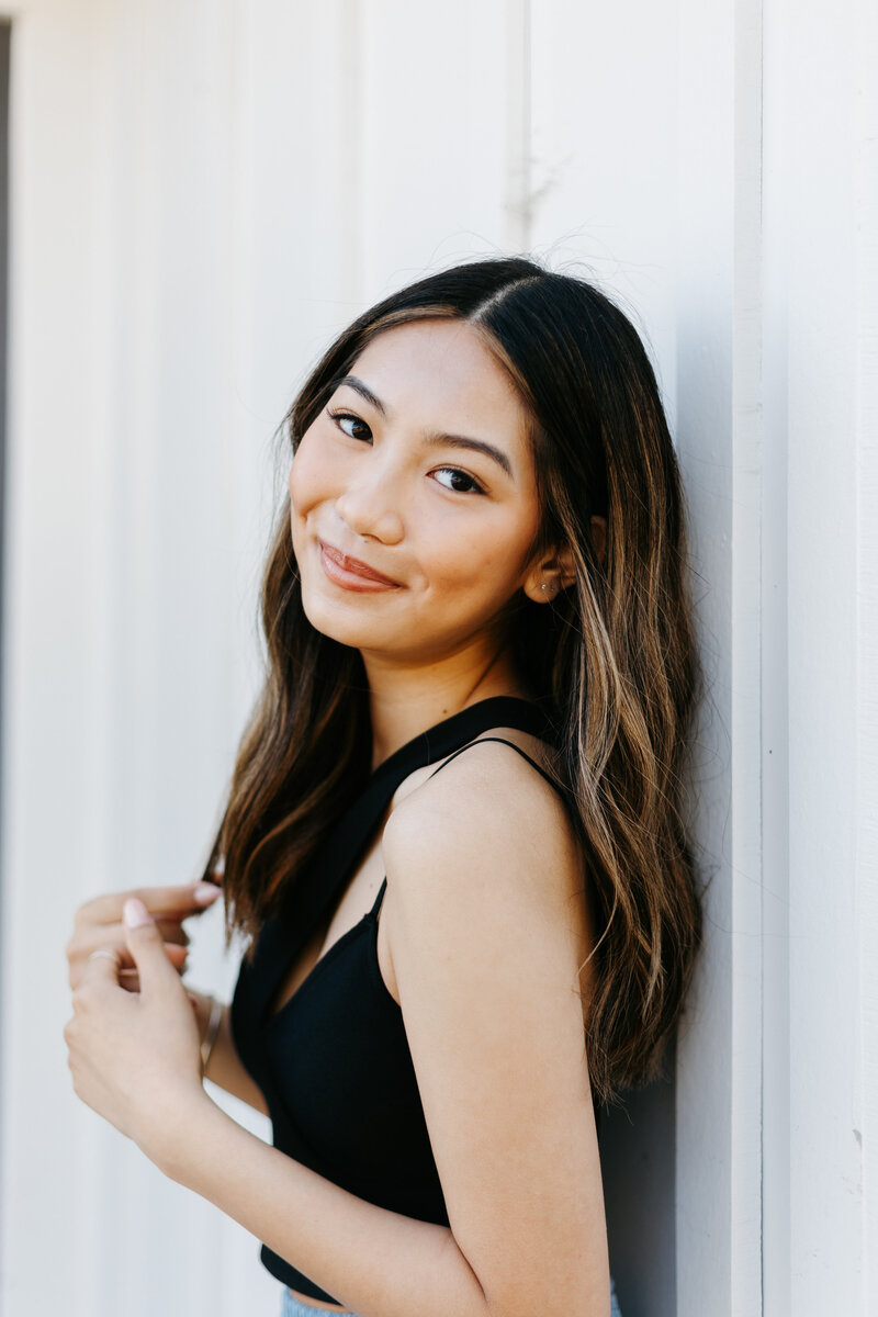 Close up portrait of a Senior girl in a black top leaning against a white wall