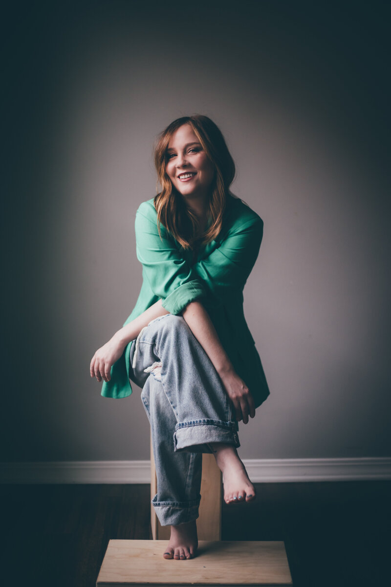 A woman with long hair wearing a green shirt and jeans sits barefoot on a wooden stool in a dimly lit room, contemplating the next steps to hire an online business manager.