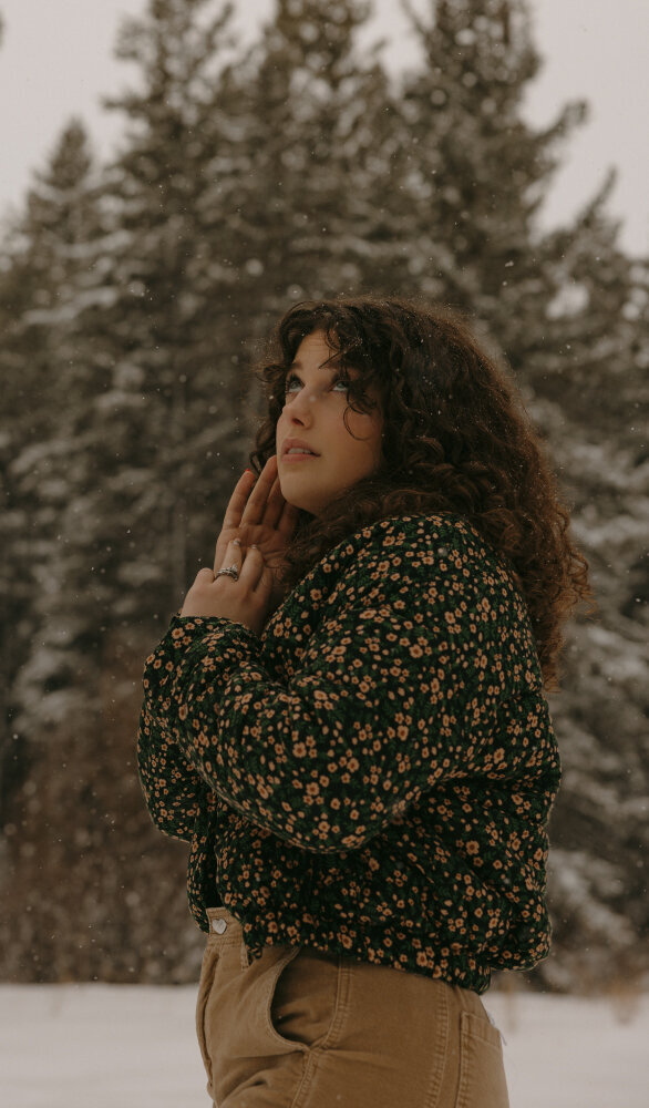 Candid, dreamy portrait of a girl outside in the snow. She has her hand lightly touching her face and she is looking up towards the sky.