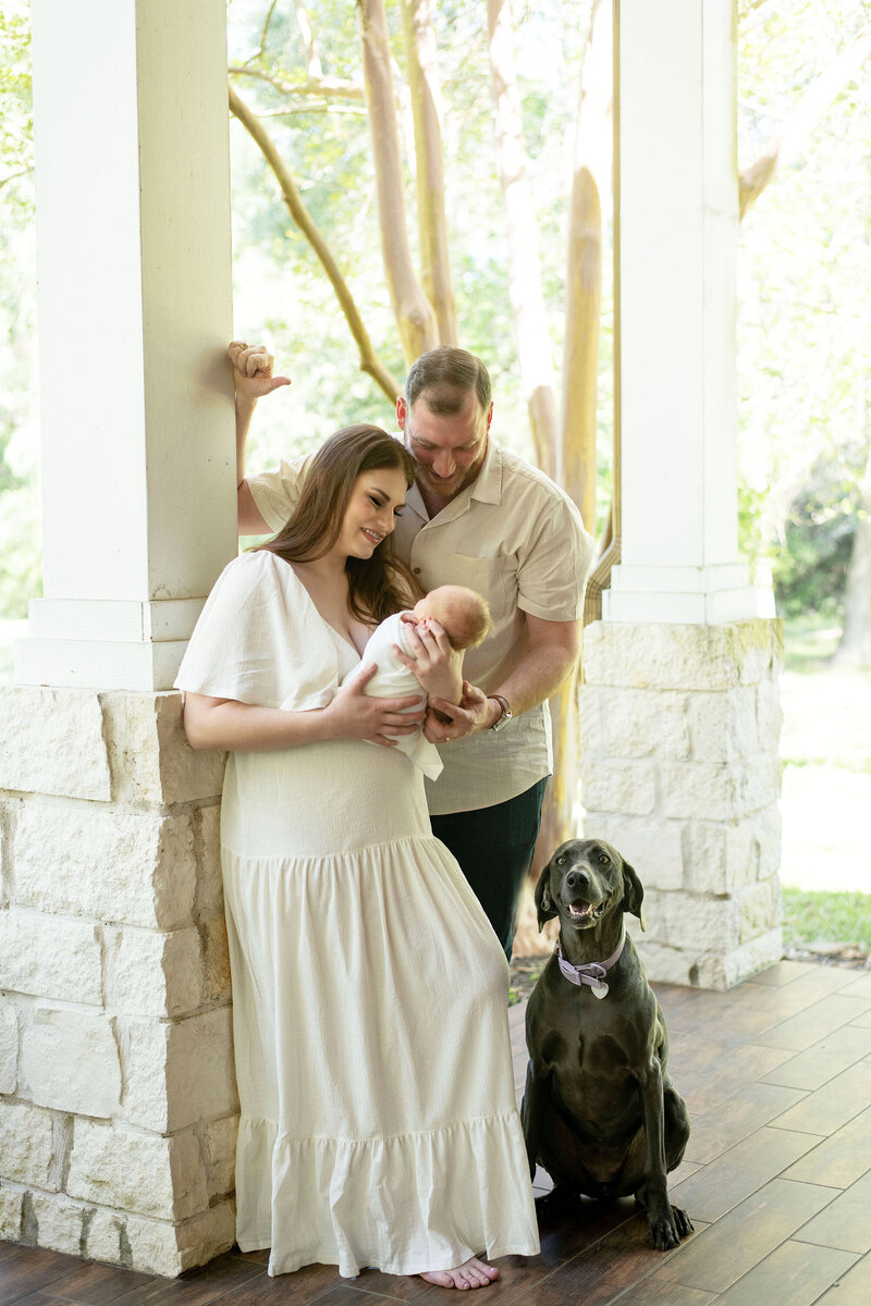 New parents hold their newborn on a cozy porch with their family dog. A warm lifestyle newborn session by Moments in Light Photography.