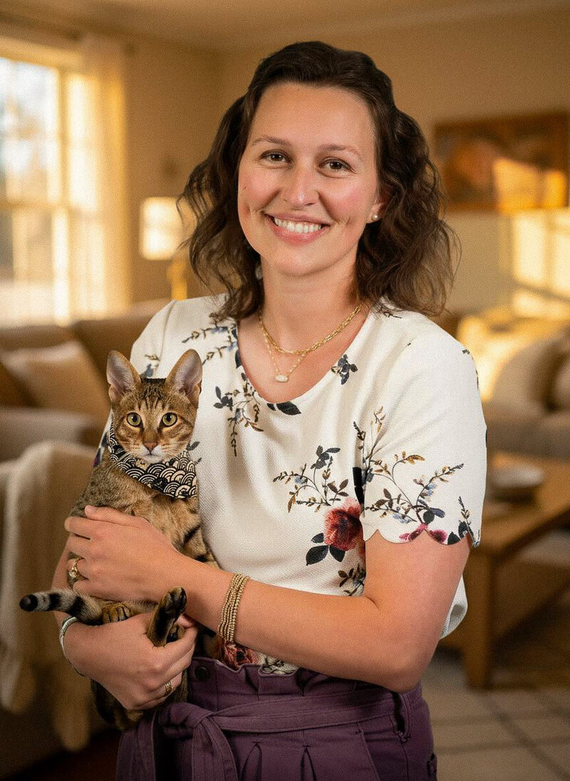A smiling woman with wavy brown hair, wearing a floral white blouse and purple pants, stands indoors holding a brown tabby cat in a patterned bandana. The warmly lit living room reflects the comfort of an eating disorder therapist in Orange County.