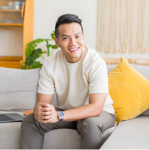 Sho Dewan, asian young man, founder and career coach at Workhap is sitting on a light grey couch with a yellow pillow. He is resting his elbows on his knees, holding his hands together and is confidently looking and smiling at the camera