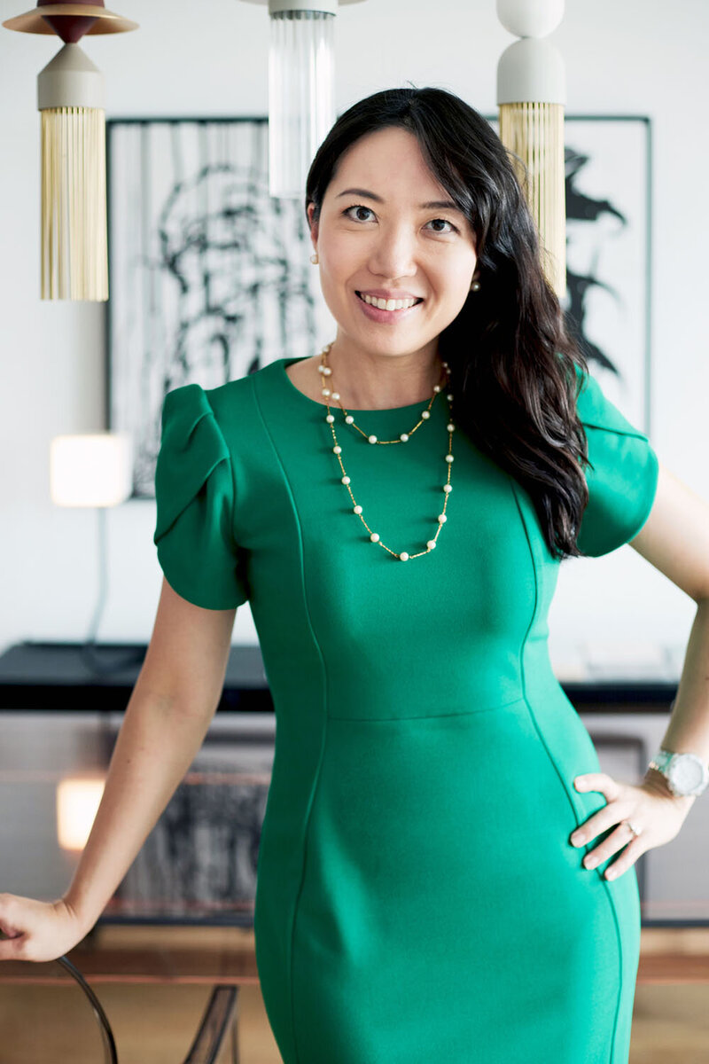 Christen in a green dress standing by a glass table in her designer studio, looking straight to camera