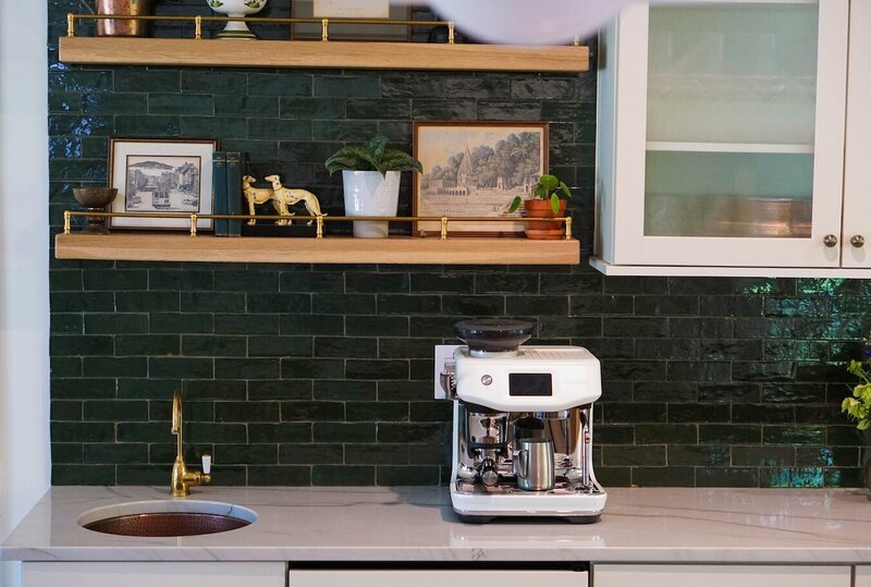Green subway tile accent wall with natural wood floating shelves, eclectic decor, small beverage sink, espresso machine, white cabinetry, and fluted glass windows.