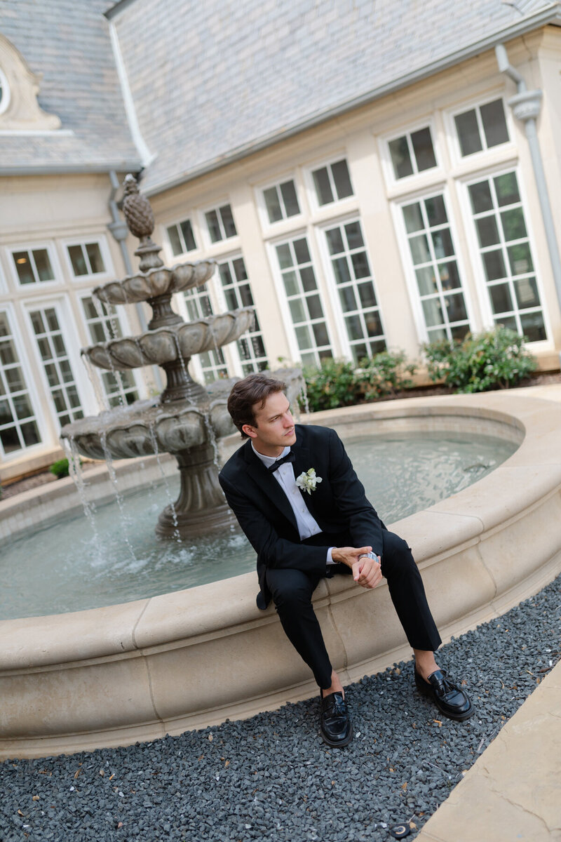 A groom in a classic black tuxedo sits contemplatively by a fountain outside a grand estate, exuding elegance and anticipation.
