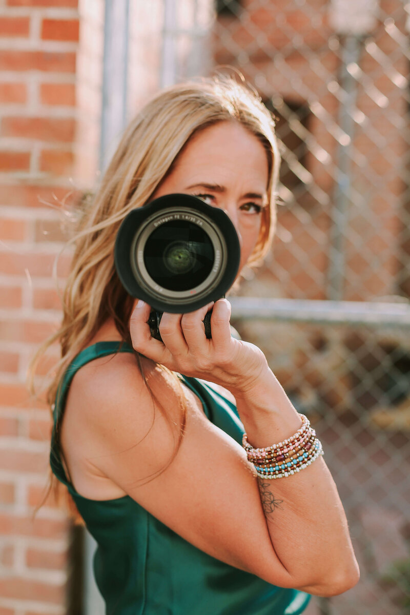 Storycatcher photographer, Kristen holding camera and looking at camera against a brick wall for a modern headshot in Kelowna.