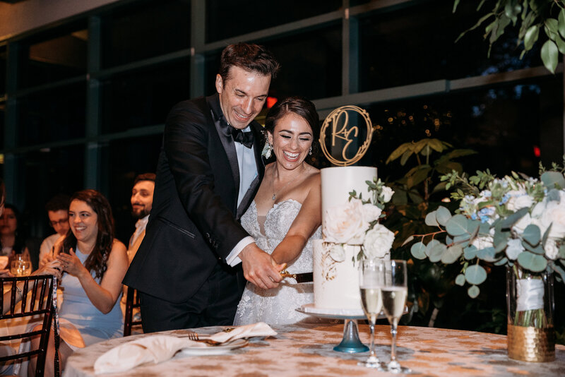 white groom in black tux and white brunette bride in strapless white gown smiling, laughing, and cutting into a 2-tier white buttercream cake with gold cake cutlery while the cake is on gold wood cake topper "R+E" sitting on a glass clear and blue cake stand with two champagne flutes on top of a table