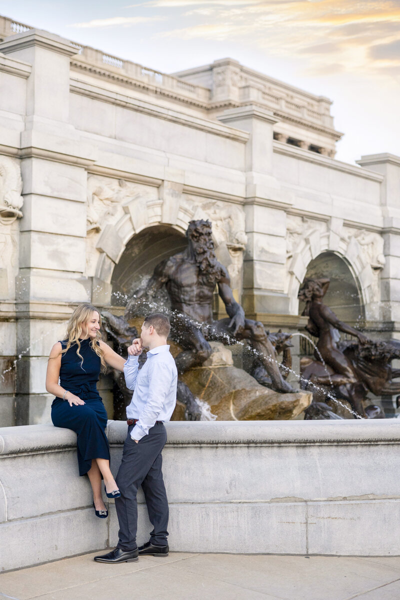 A woman in a navy dress sits on the edge of a fountain with stone sculptures, while a man in a light shirt and dark pants stands beside her, holding her hand. They are both smiling against the backdrop of an ornate building and a sunset sky.
