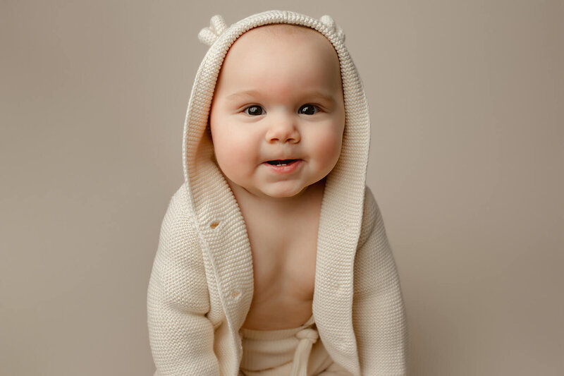a 6 month old sitting on a white cloth looking and smiling at the camera
