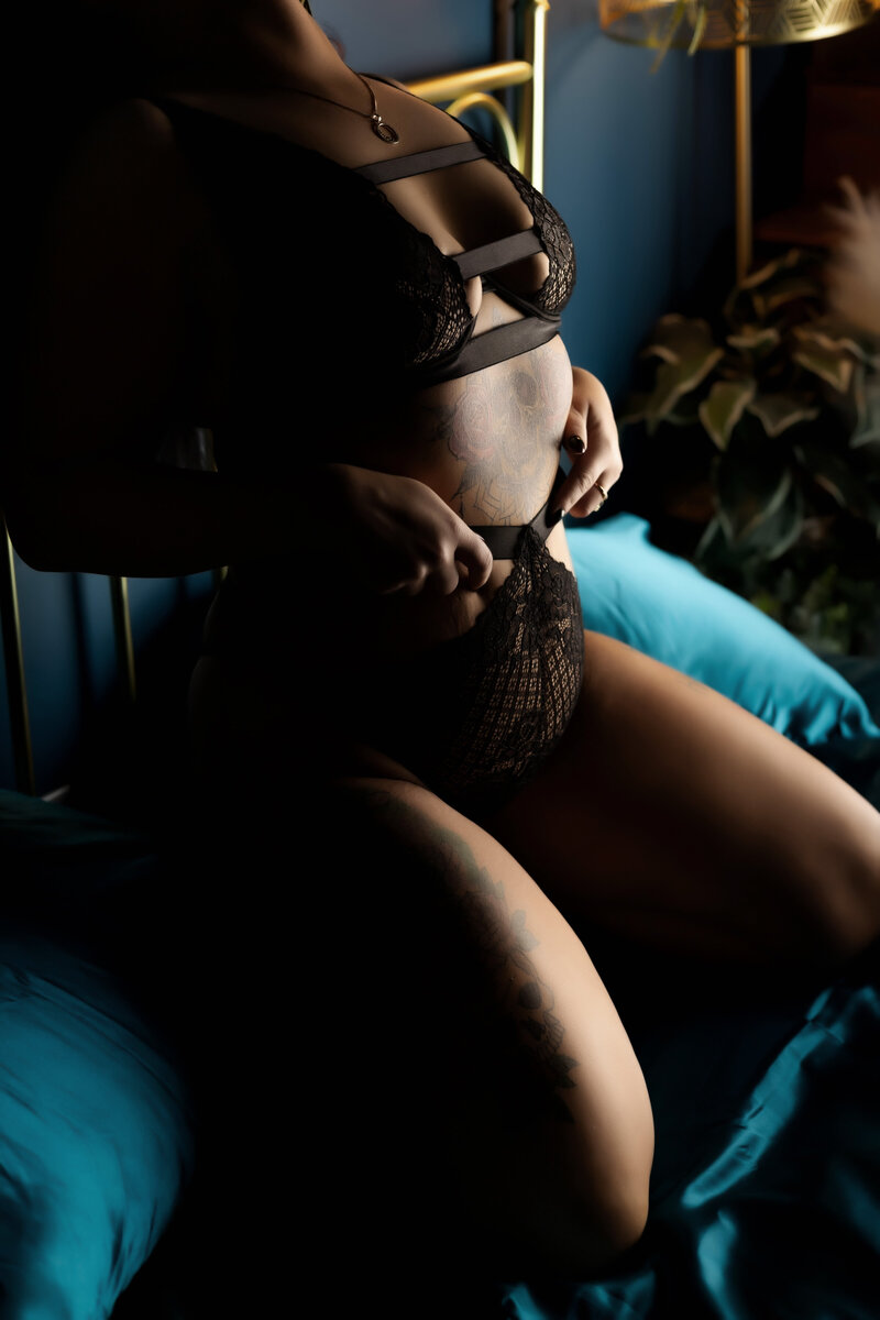Close-up boudoir shot of client sitting on her heels with knees tucked, leaning against a headboard while pulling up on the waistband of her panties — favorite pose photographed by Joclyn in Cedar Rapids, Iowa