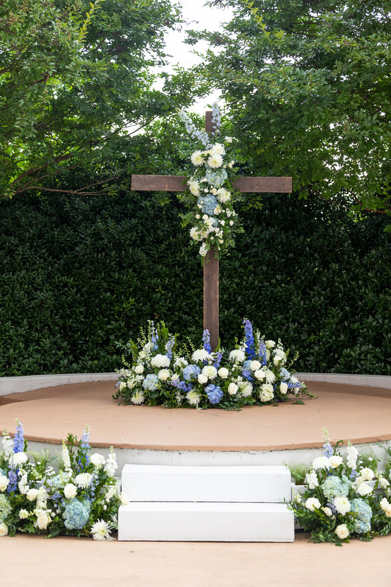 Spring wedding at WinMock at Kinderton, designed by Charming Carolina — wooden cross with a white chiffon drape and matching dusty blue florals for the ceremony backdrop.