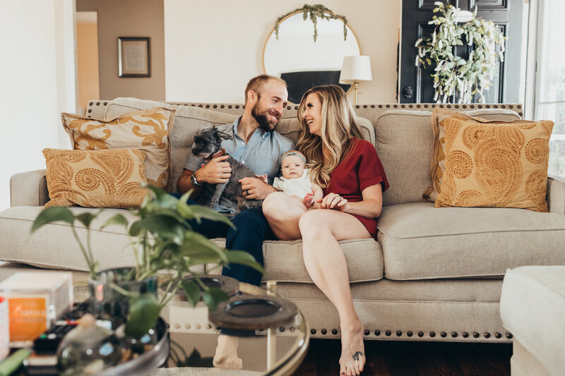 A family of three sit on the couch laughing together while holding their dog in their La Mesa home.