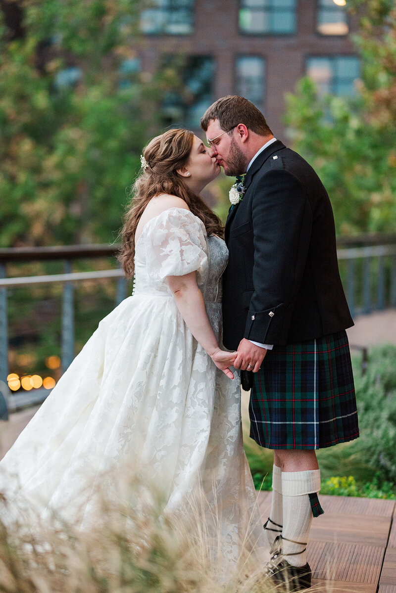 jewish bride kissing and holding hands with scottish groom on a terrace with a greenery backdrop