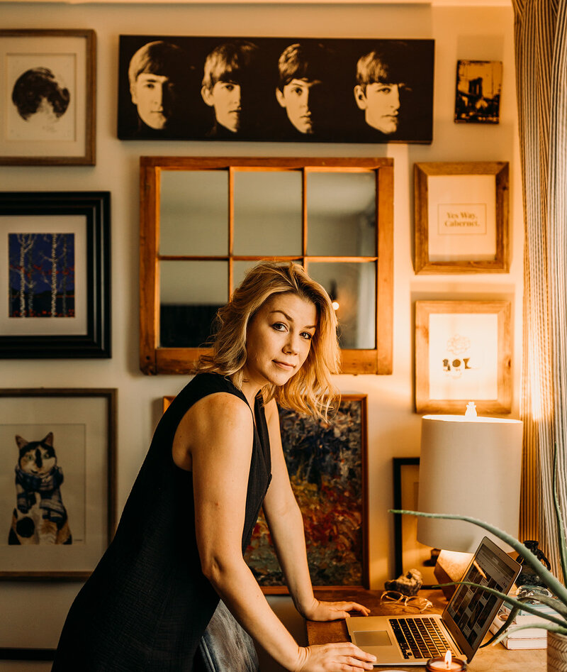 Lauren glances towards the camera as she leans on her work desk. Her laptop is open, and she's standing in front of a gallery wall.