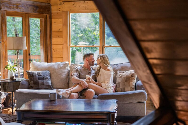 A bride and groom sit on a couch at a cozy cabin in the woods enjoying coffee. The bride has her legs over the grooms and he is gently holding them. They are looking at each other smiling lovingly. 