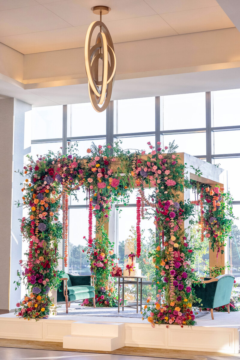 A beautifully decorated ceremonial mandap adorned with vibrant flowers in shades of pink, purple, orange, and green foliage. Two green chairs and a small table are placed beneath it. Large windows in the background allow natural light to flood the space.