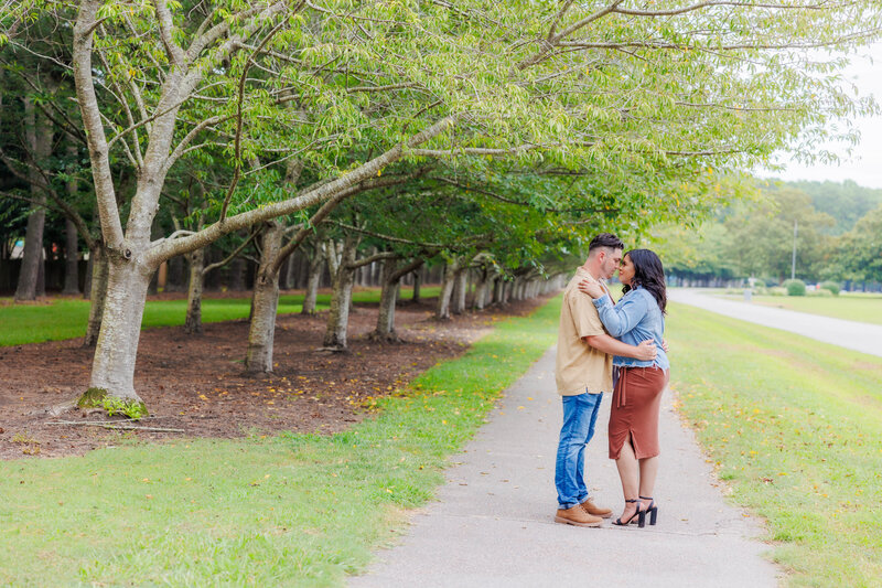 Fall in Virginia at an Engagement Session in Red Mill Park.  Bride and Groom to be are dressed in neutral tans, browns and denim.  Couple is looking deeply into each others eyes as they are facing each other and embracing.  Picturesque location provides a lined path with trees.  Virginia Wedding Photography by Angela Foushee.