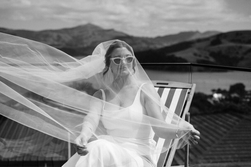 black and white shot of bride with veil over face in akaroa new zealand on striped deck chair overlooking ocean