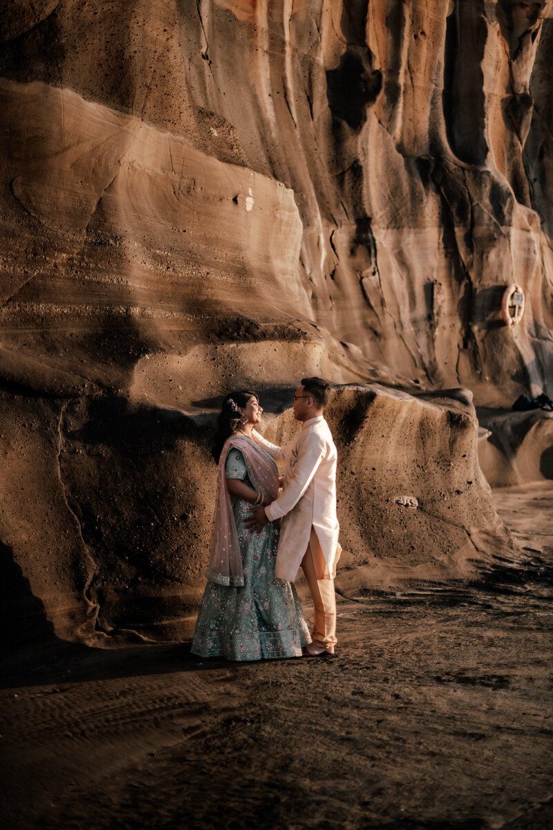 Couple wearing traditional Indian wedding attire posing against the dramatic cliffs at Muriwai Beach, Auckland, captured by Zanthe Vorsatz Photography.