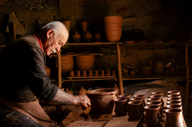 Elderly potter with silver hair wearing a brown shirt, khaki apron, and red scarf, bending over a bench molding a clay bowl in a dark workshop filled with pottery, symbolizing God as the potter shaping lives as described in Isaiah 64:8.