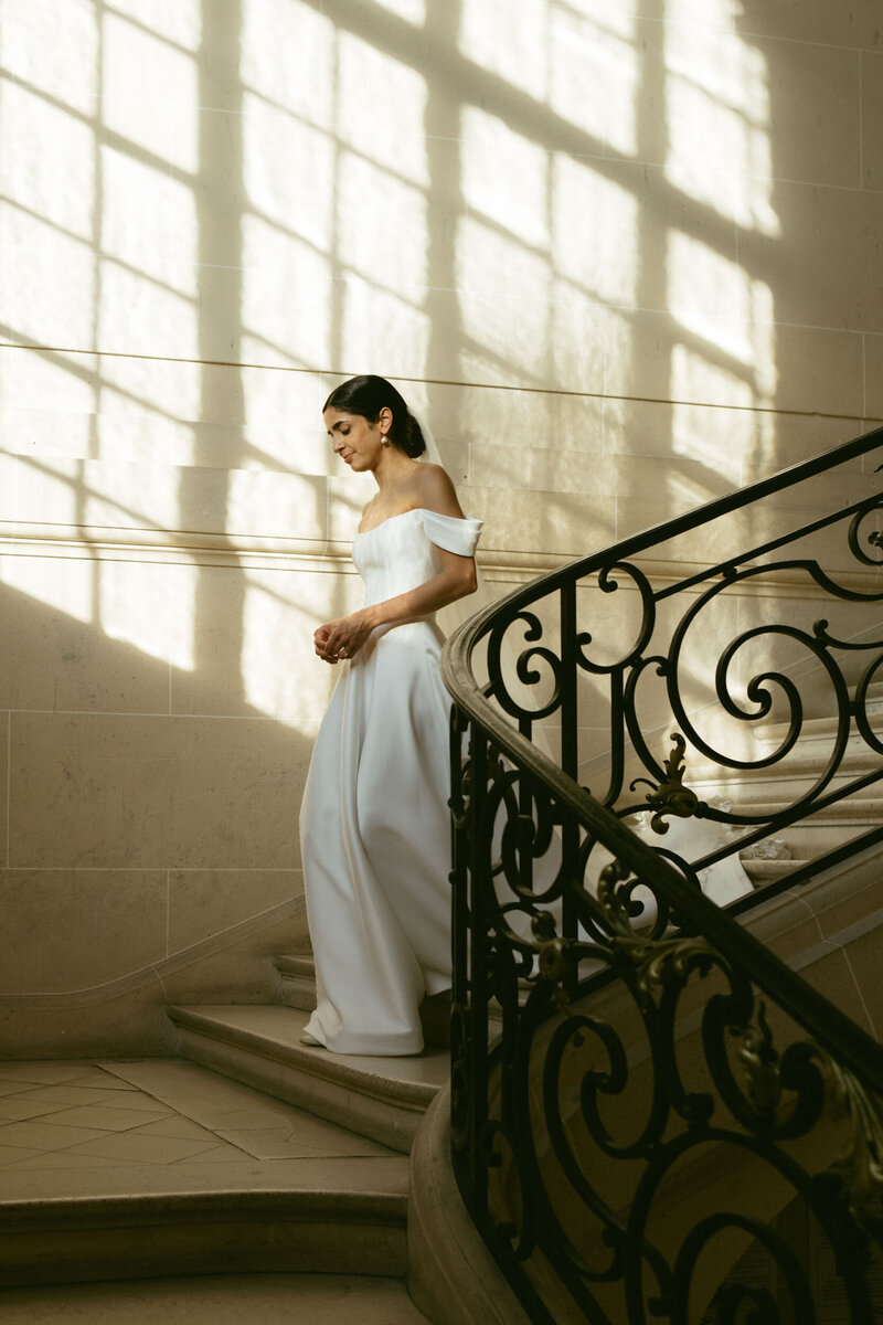 A bride walks down the staircase of a historic French Chateau with window light shadows, shot with a film aesthetic by Stacey Vandas Photography.