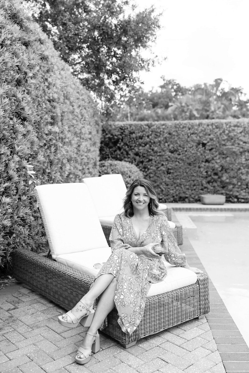 woman sitting on lounge chair with wine by a pool surrounded by hedges