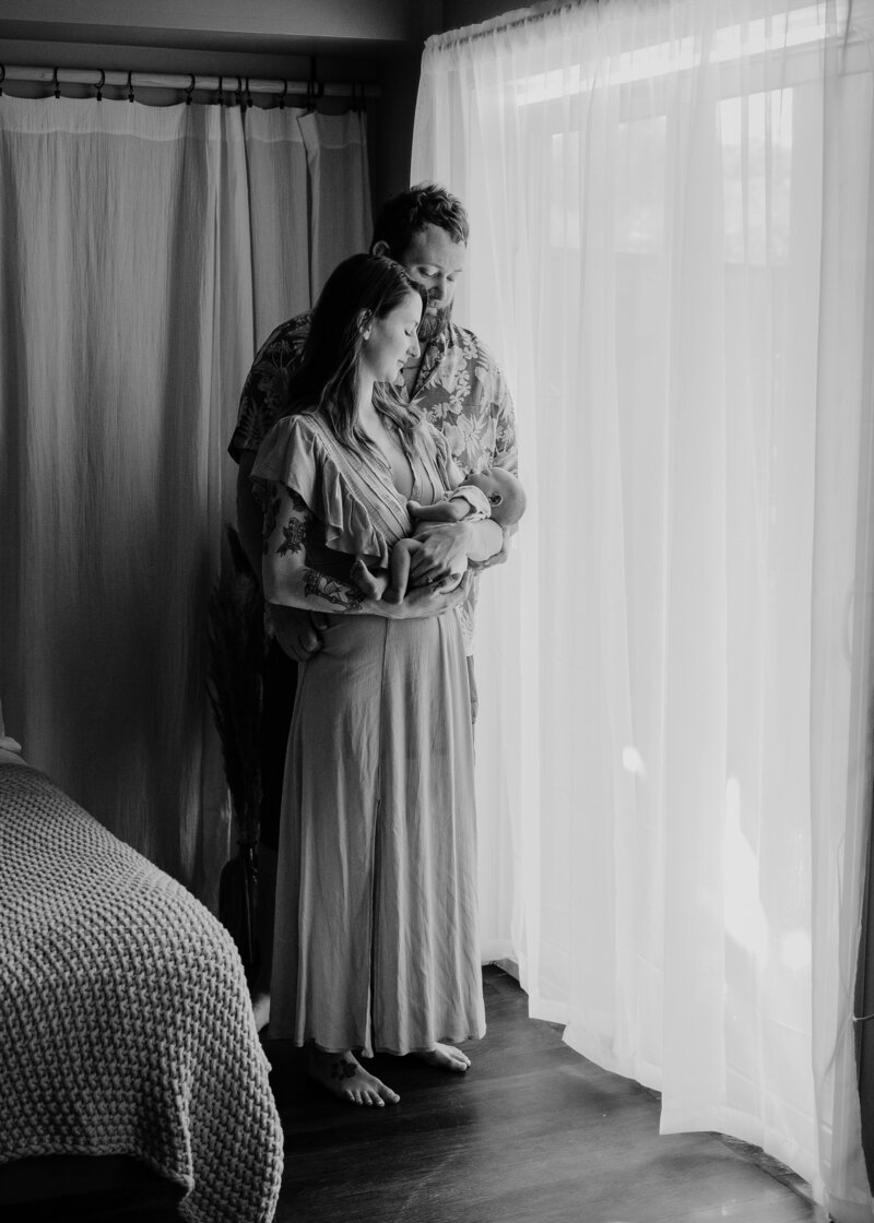 Black and white newborn photo of mom standing and holding baby next to studio window with dad standing behind her while looking at baby in Severn Maryland studio photographed by Bethany Simms Photography