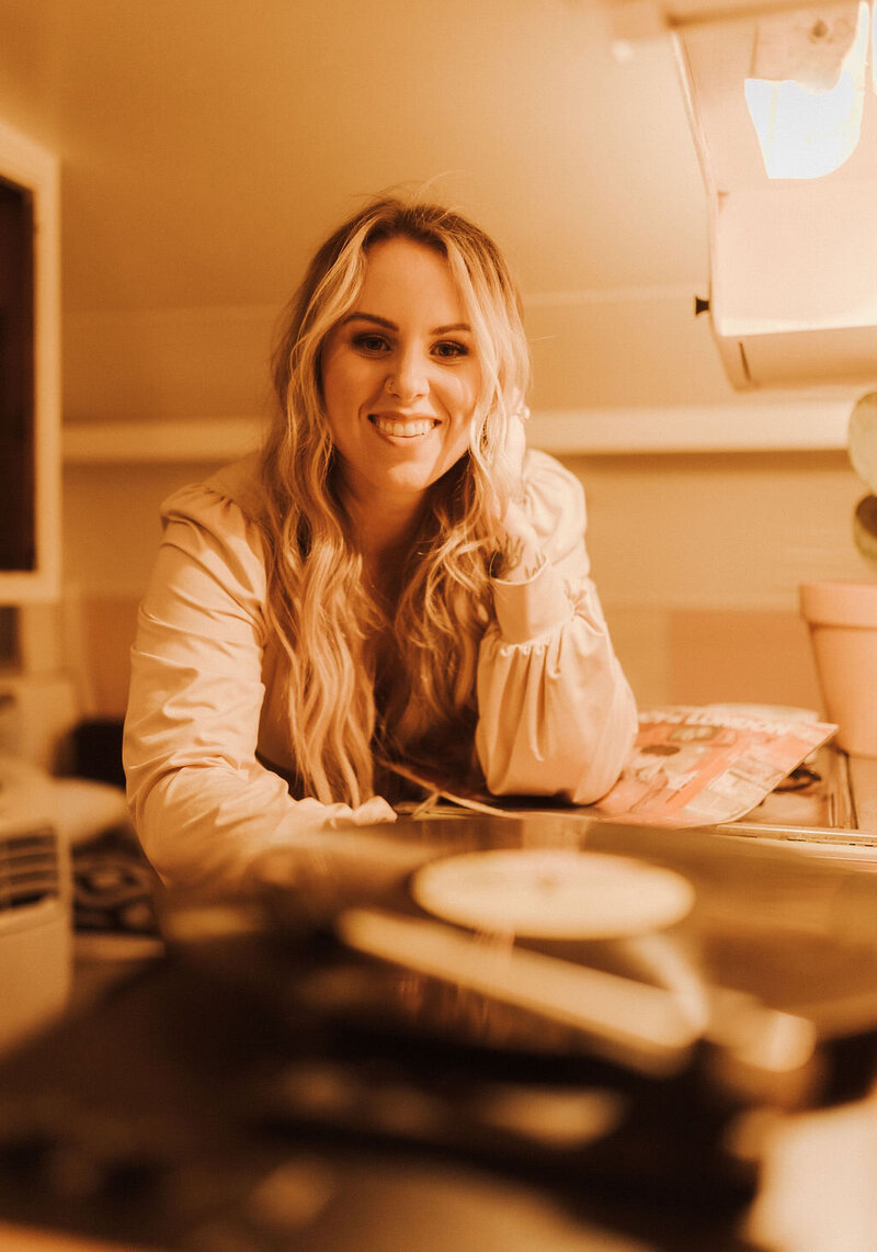 Specialist in seamless real hair extensions is sitting on a beige sofa. Her hair is blonde, long, and wavy. She is wearing an elegant white blouse.