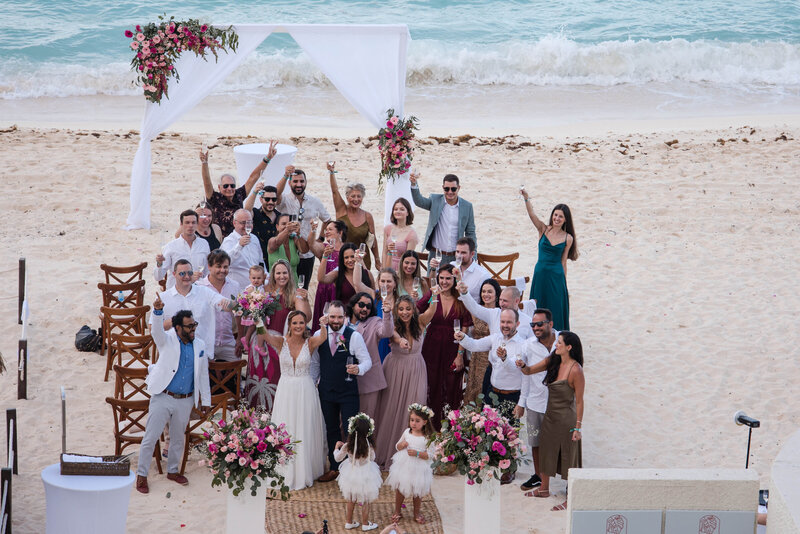 Microwedding of bride and groom and their guest raising their glass toward the photographer celebrating t a beatufil beach with a colorul arch against turquoise water and a colorful splash of an arch with spray of pink, burgundy bouquets.