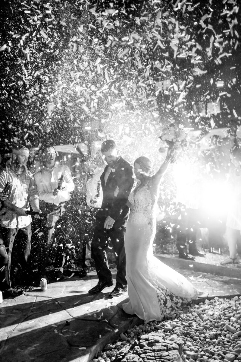 A newlywed couple walks hand in hand through a shower of confetti, illuminated by dramatic lighting during their grand exit