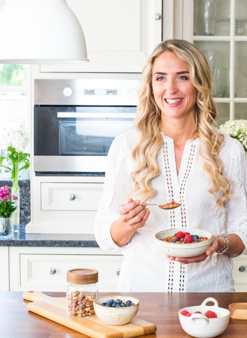 Charlotte Grand, founder of The Fertility Kitchen, smiling in a bright, inviting kitchen while holding a bowl of granola topped with fresh berries. She wears a white embroidered blouse and stands behind a wooden countertop adorned with nourishing ingredients like nuts and fruit, embodying her passion for fertility-focused nutrition.