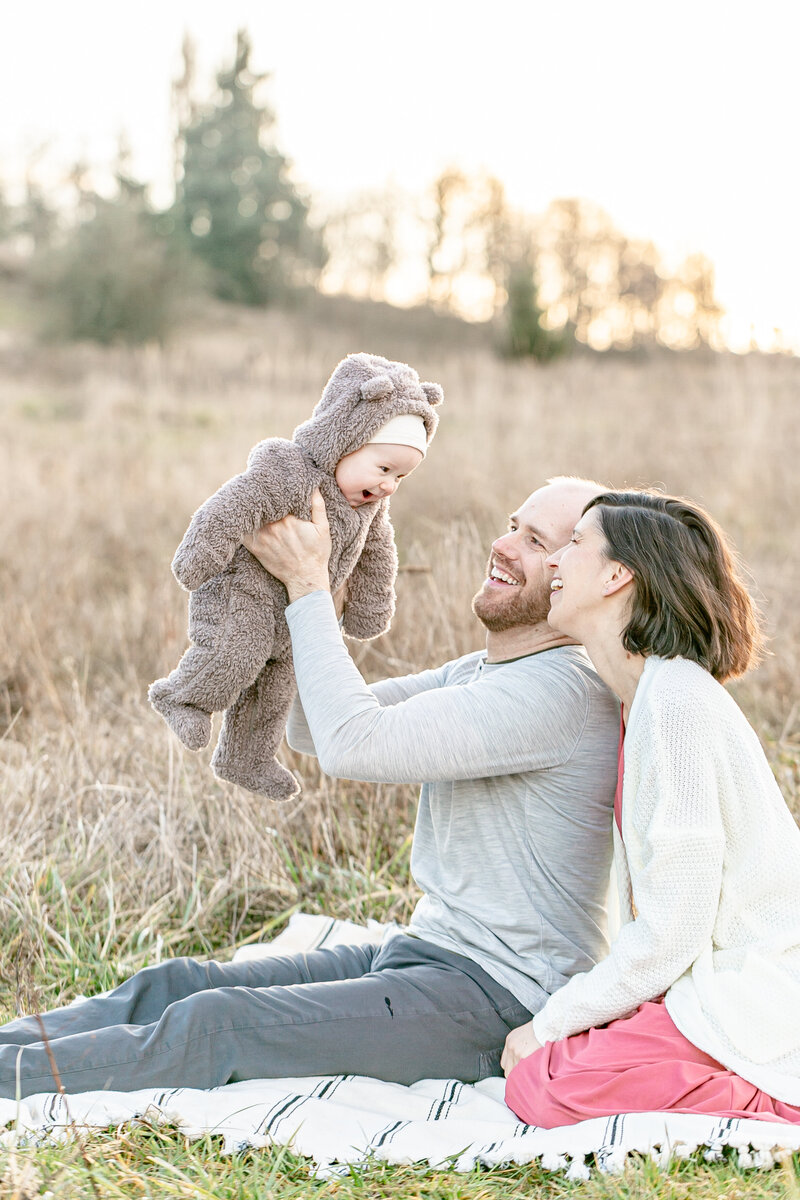 Parents sitting on a blanket out in nature with six month old baby in a little tan fleece bear suit all laughing with each other