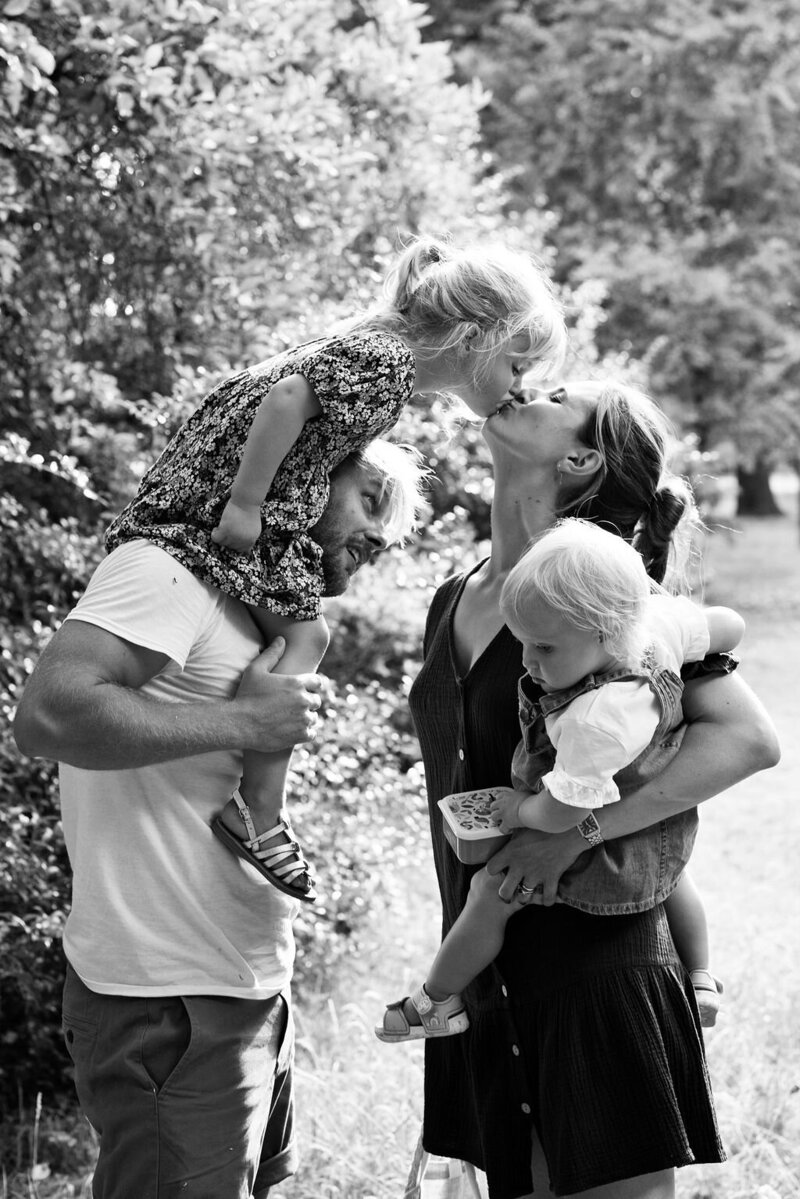 Mother kissing little girl as she sits on her father's shoulders during a photo shoot in Bushy Park