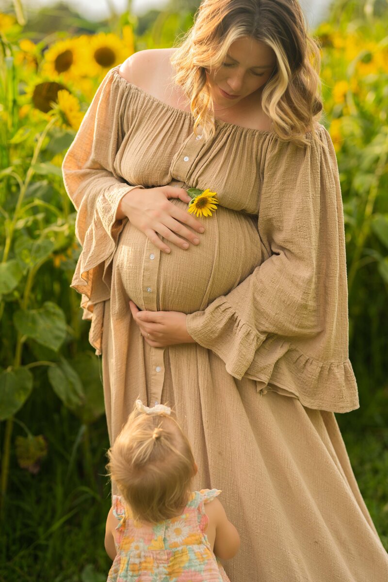pregnant mom in medina sunflower field holding belly and looking fown at her toddler daughter