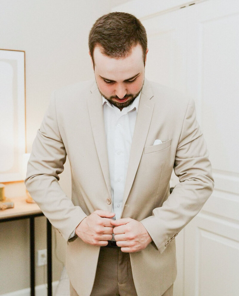A groom pulls his light tan suite jacket tight in the crazy gorgeous bridal suite.