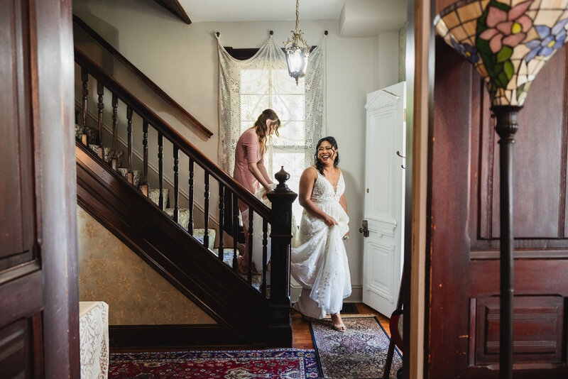 A person in a wedding dress walking down the stairs while someone holds their train behind them