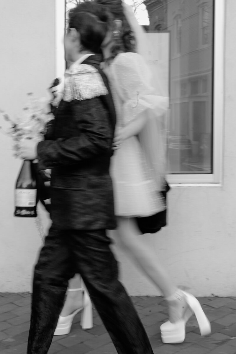 Blurred black and white image of nonbinary wedding couple walking the sidewalk in Georgetown DC while holding a bouquet and champagne bottle