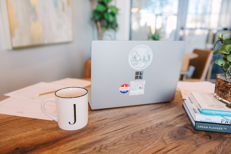 Lap top sitting on a table next to a cup of coffee with the initial "J"