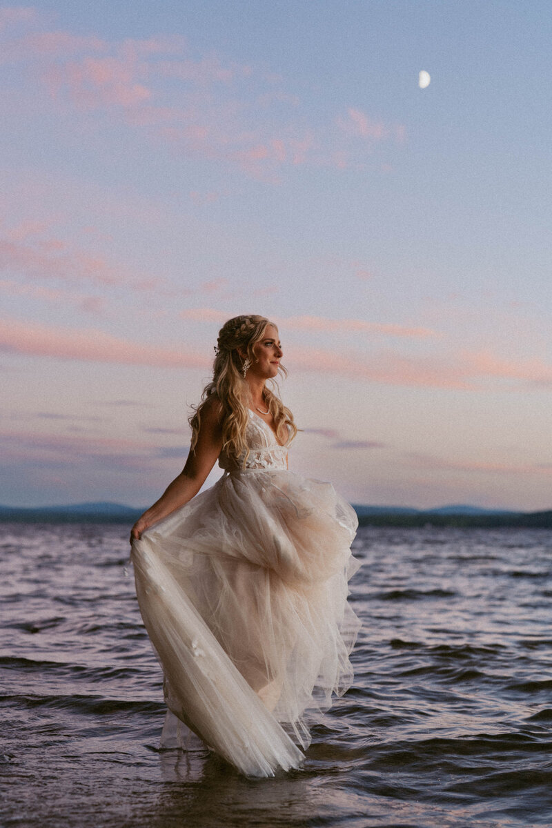 bride standing in a lake, twirling her wedding dress against a pastel blue and pink backdrop