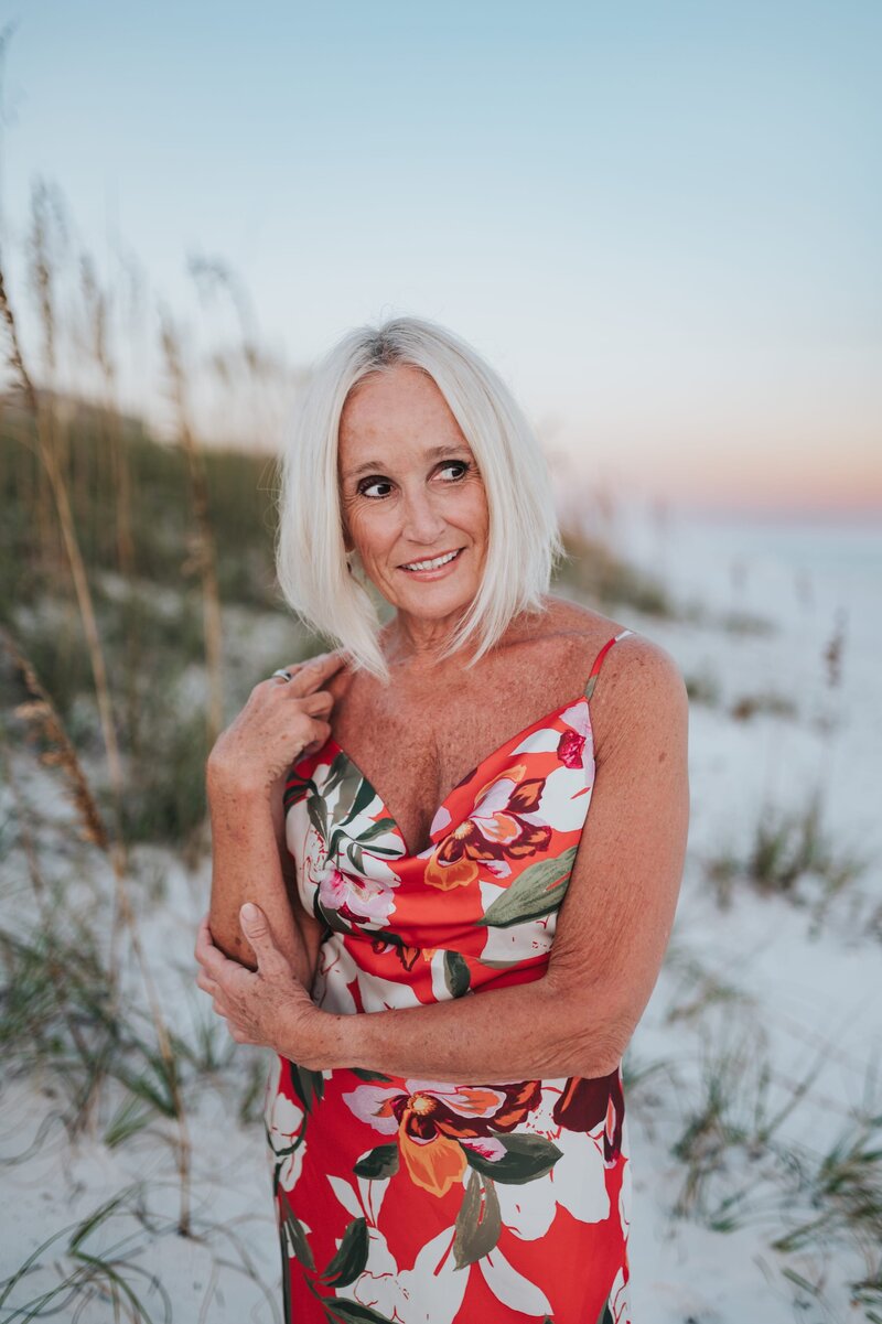 Woman with a white bob smiles off camera while wearing a vibrant, floral dress as she stands on a Florida beach at sunset,