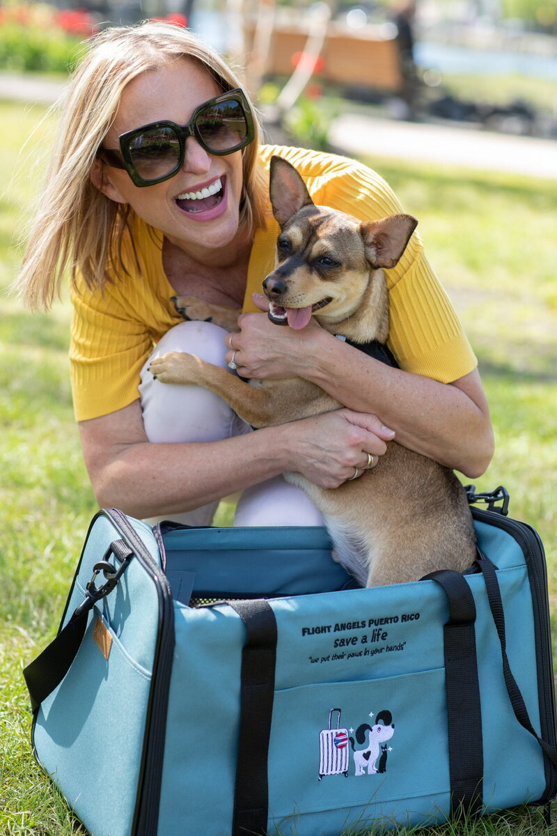 Woman in yellow sweater kneeling in park on sunny day takes chihuahua out of blue carrying case 