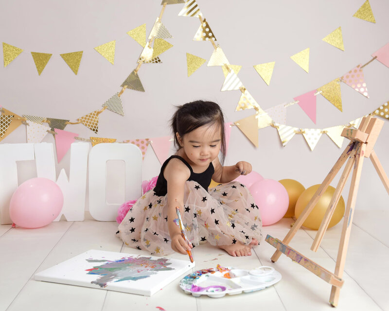 A toddler painting on a canvas in a creative two-year milestone photo shoot, featuring golden and pastel bunting, balloons, and an art-themed setup.